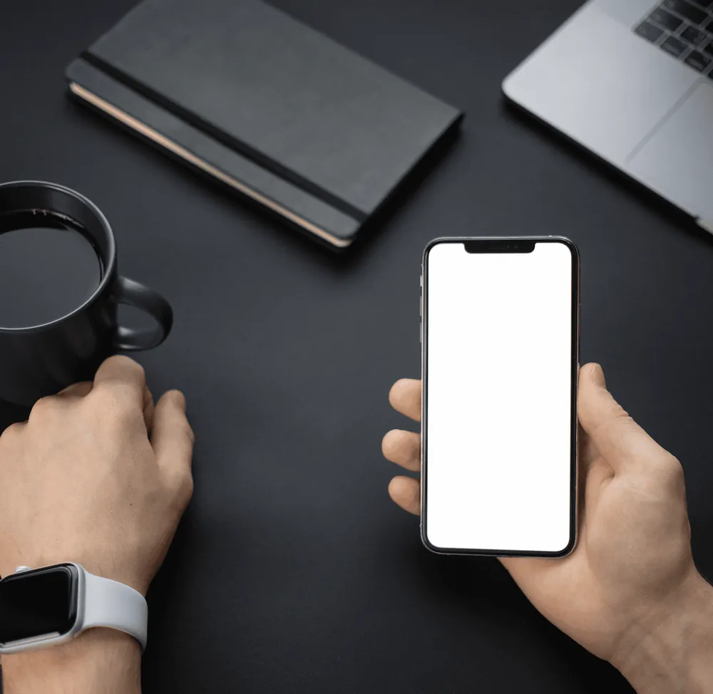 Hands holding a phone with a blank screen above a dark desk with a notebook and a mug