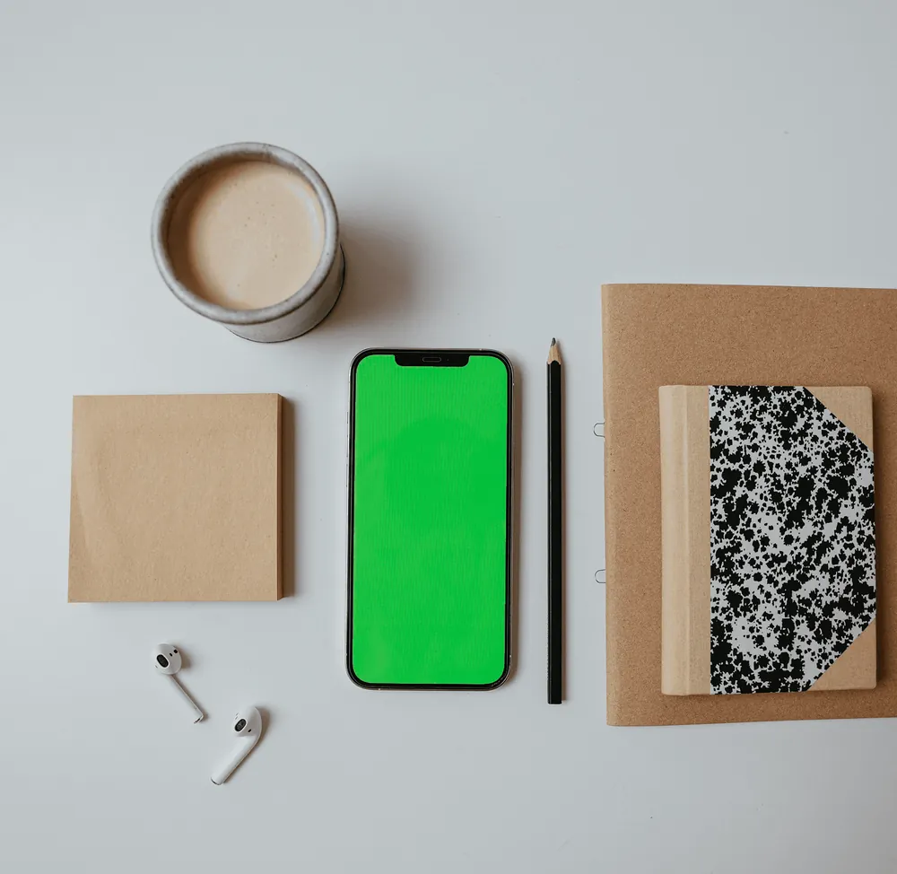 A green-screen phone laid out beside notebooks, earbuds and a cup of coffee