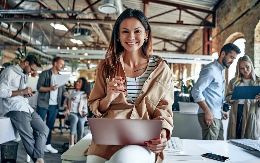 A smiling woman with a laptop in a busy office