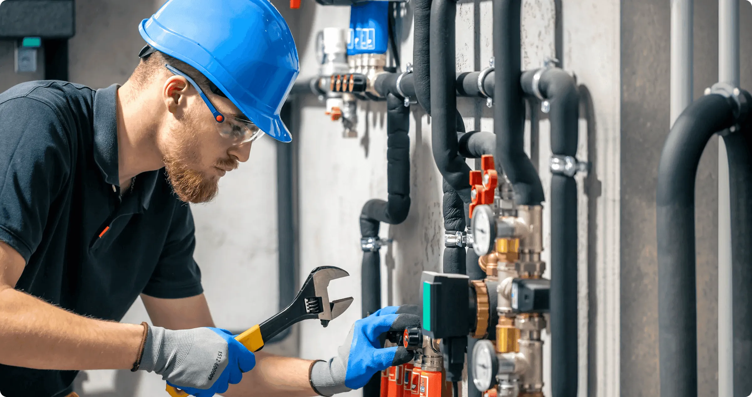 A plumber in a blue hard hat adjusting valves on a heating system with a wrench