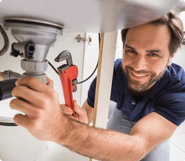 A smiling plumber tightening a sink drain with a red pipe wrench