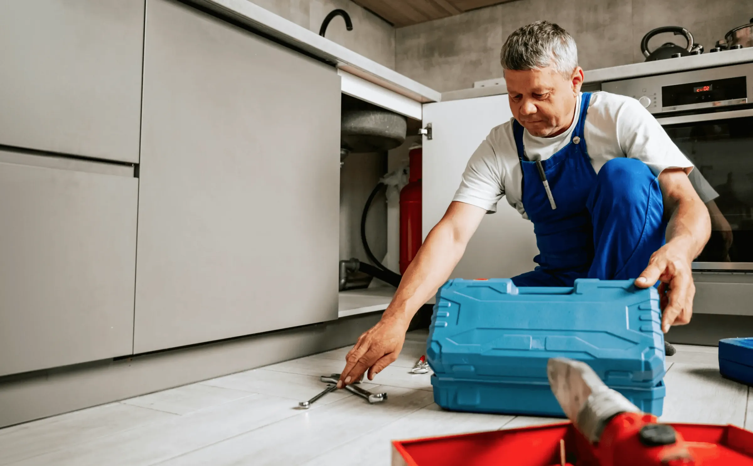 A plumber in blue overalls kneeling by an open kitchen cabinet with his toolbox