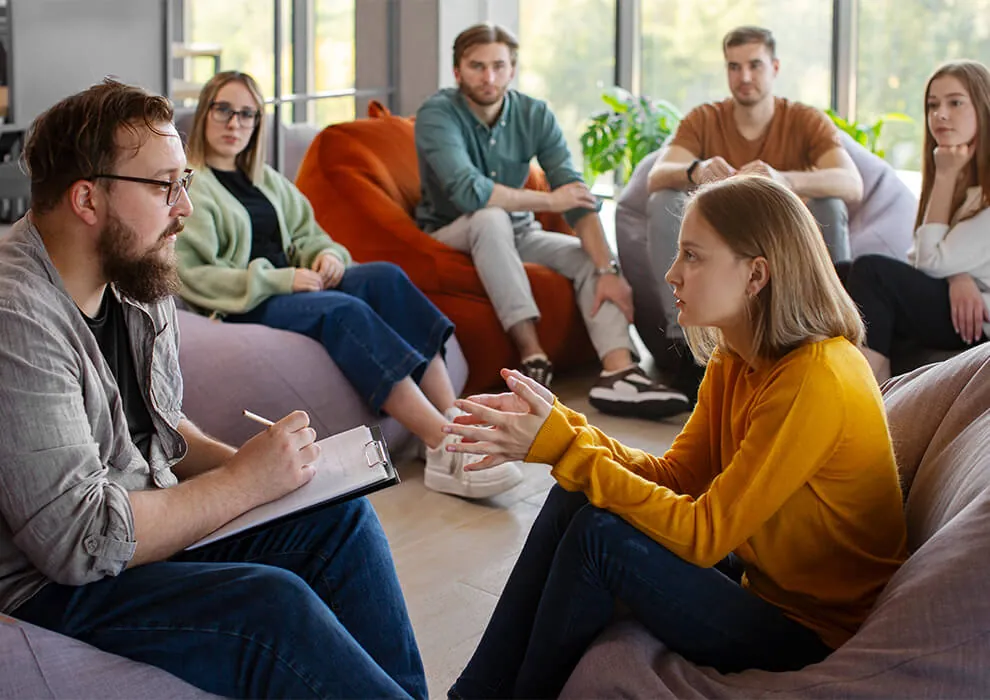 A therapist taking notes while a young woman speaks in a group session