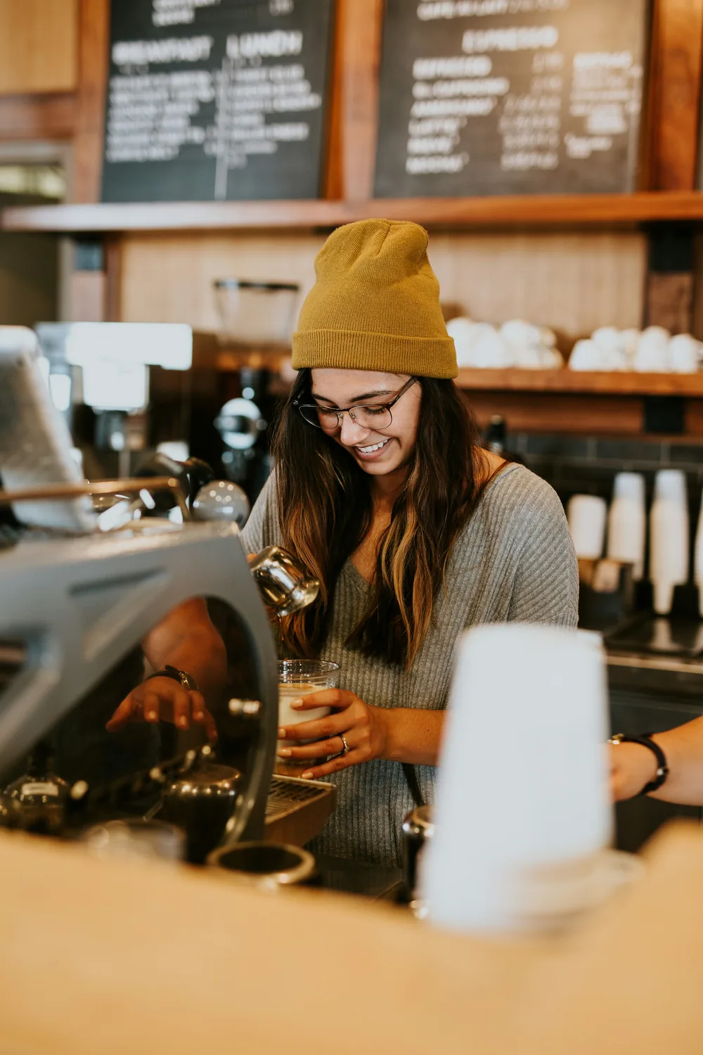 A smiling barista in a yellow beanie making coffee behind the counter
