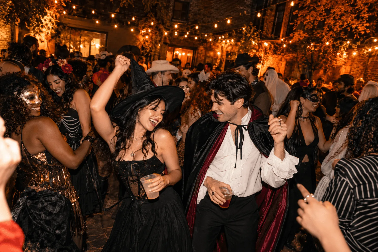 Costumed adult guests dancing together beneath amber courtyard string lights