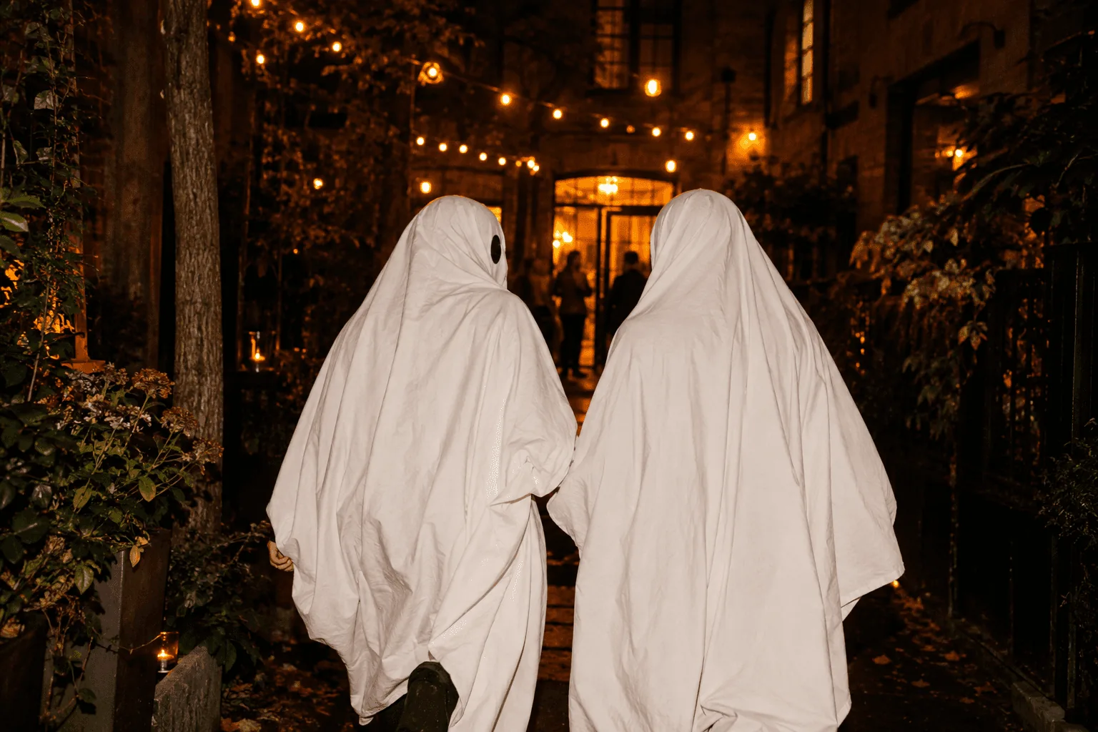 Two friends in sheet ghost costumes walking toward a warmly lit doorway after the party