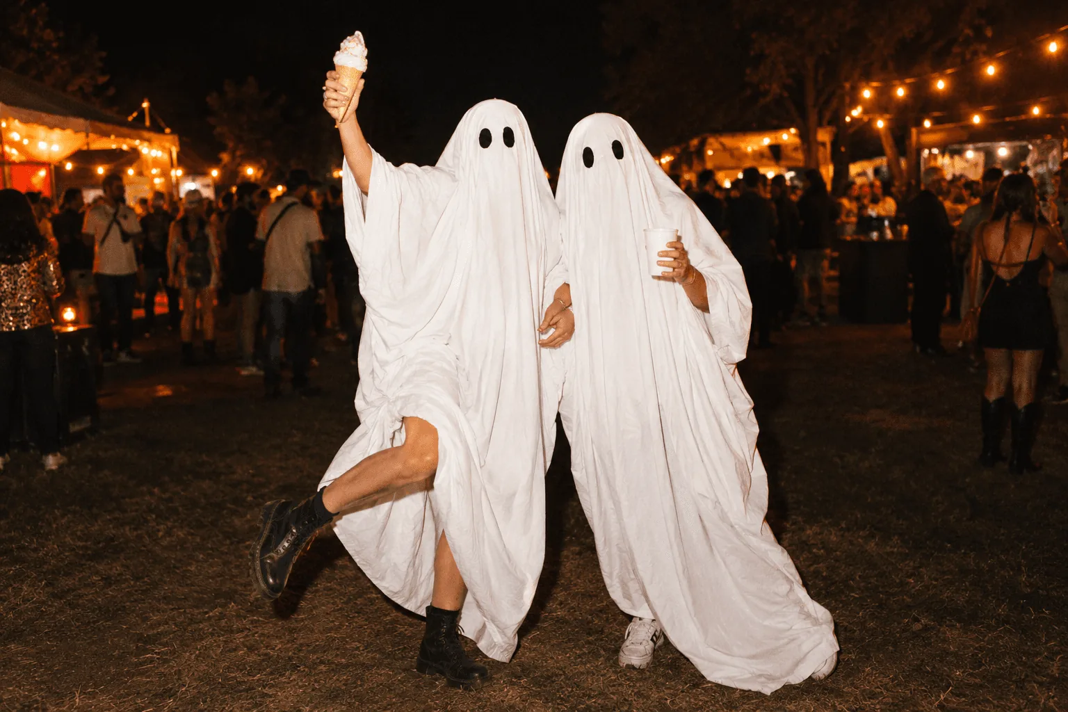 Two friends in white sheet ghost costumes enjoying ice cream at a warmly lit Halloween party