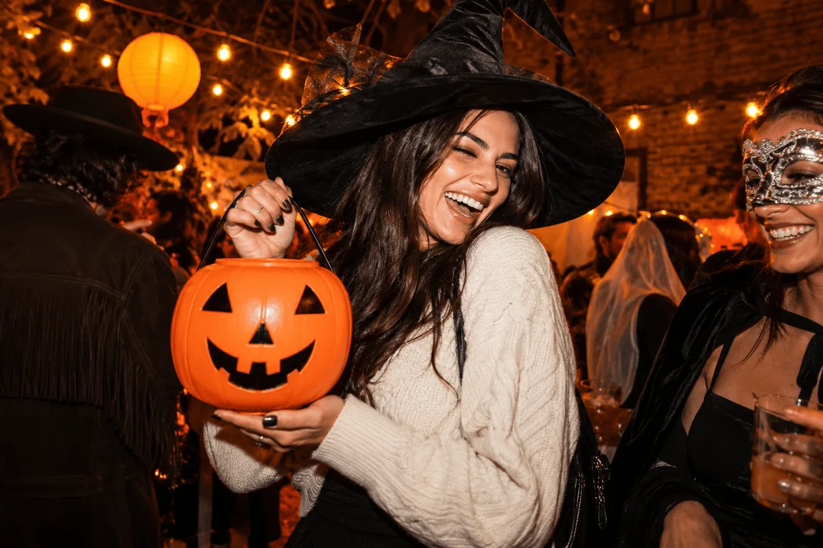 A guest in a handmade witch hat holding a pumpkin candy bucket beside a masked friend