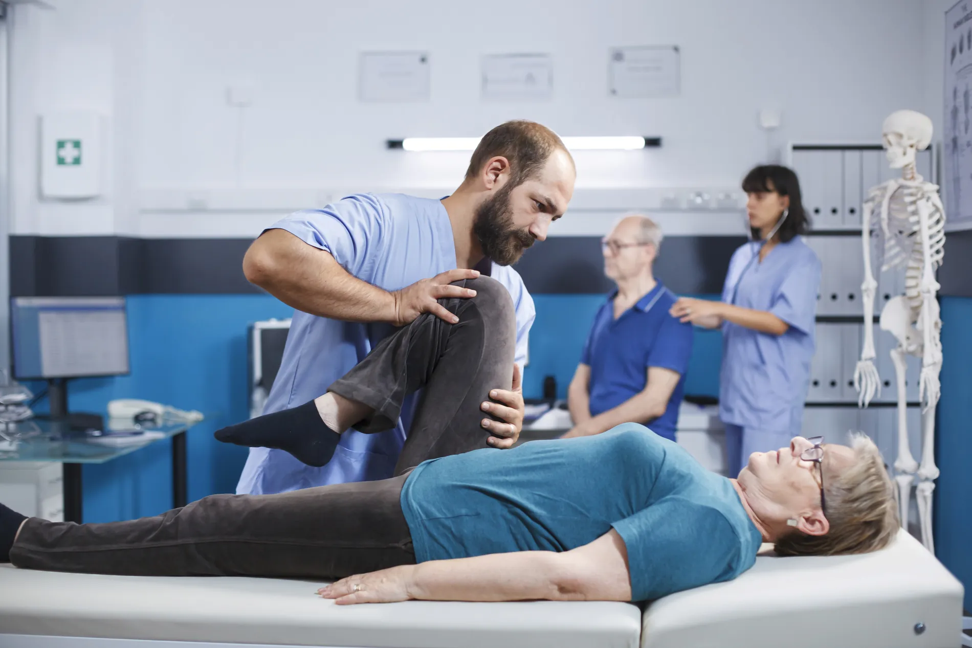 A chiropractor bending the knee of a patient lying on the treatment table while colleagues work nearby