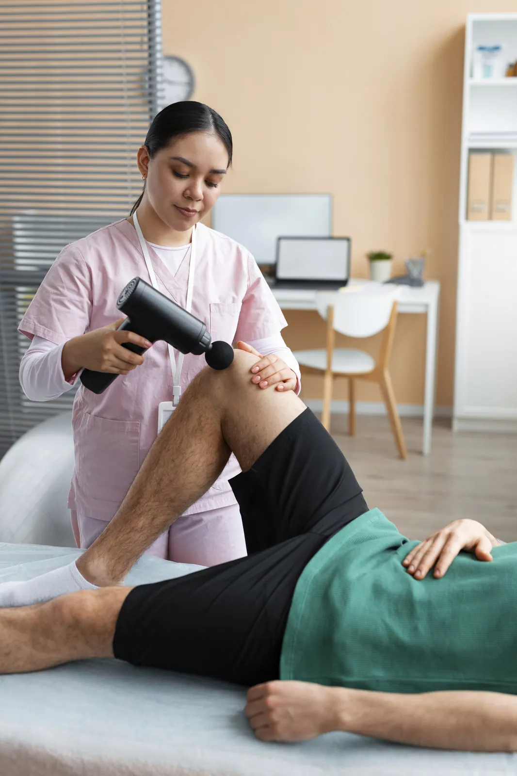 A physiotherapist using a massage gun on the knee of a patient lying on the treatment table
