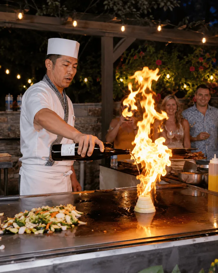 A chef sending up a tall flame from the grill at an evening garden party