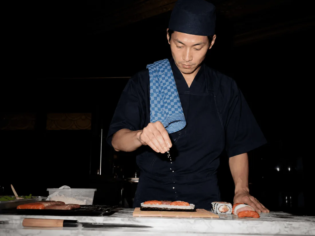 A chef in a dark jacket plating sushi under warm light
