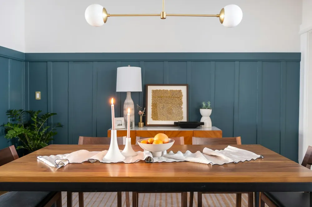 A dining room with blue panelled walls, a wooden table and a brass wall light