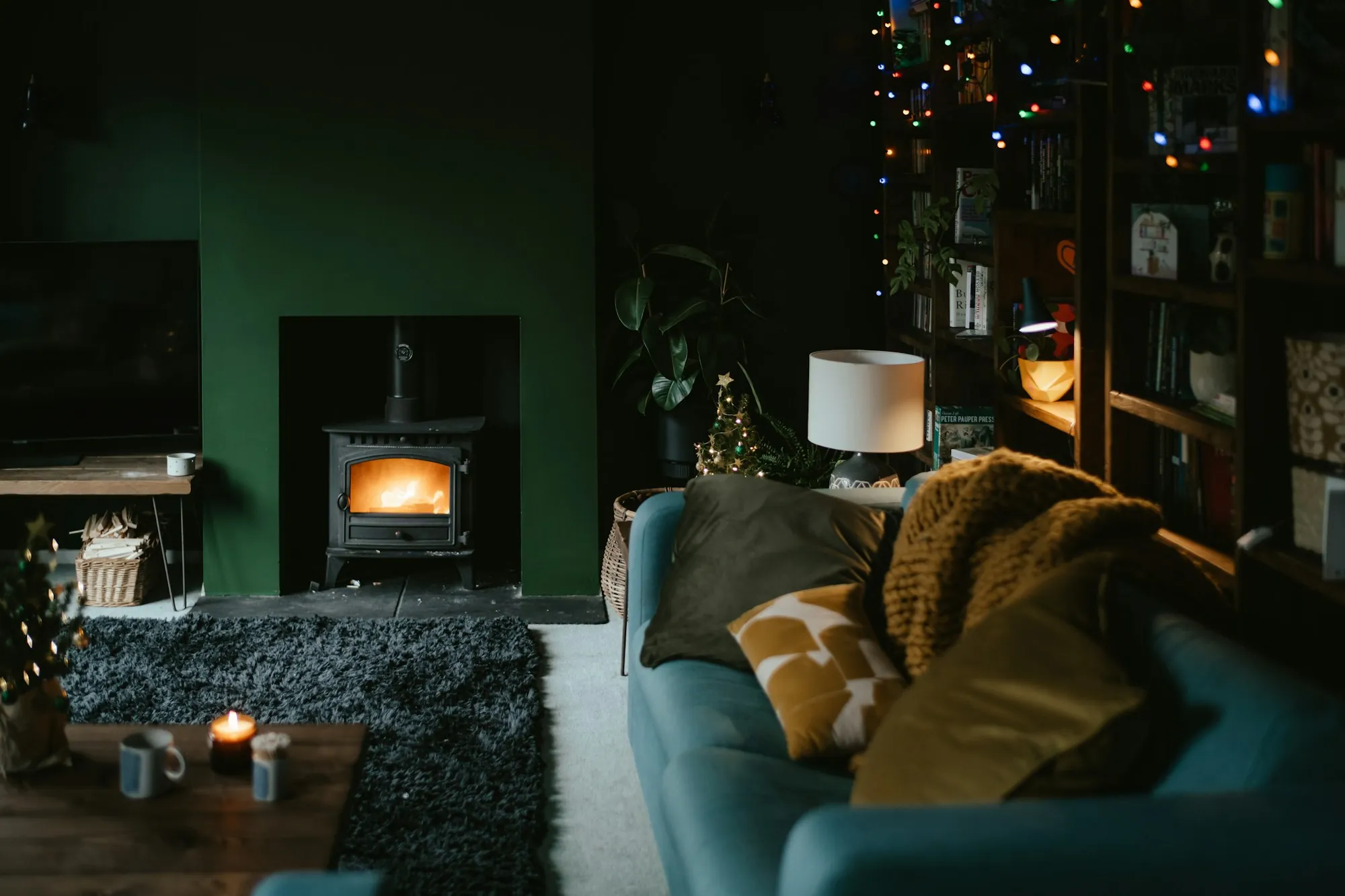 A deep green living room with a wood burner, a teal sofa and warm lamplight