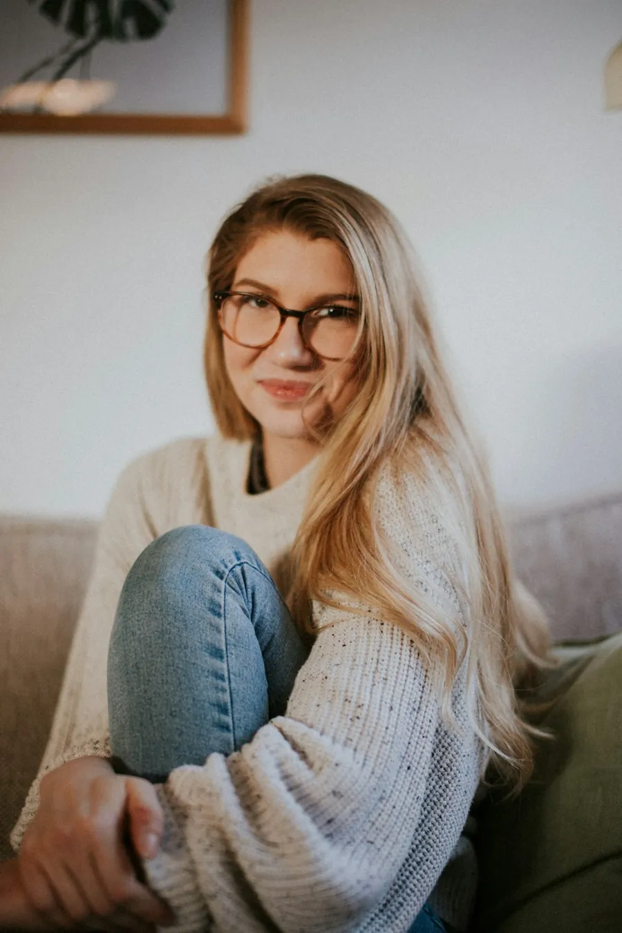Clare, smiling in a cream jumper and glasses, sitting on a sofa