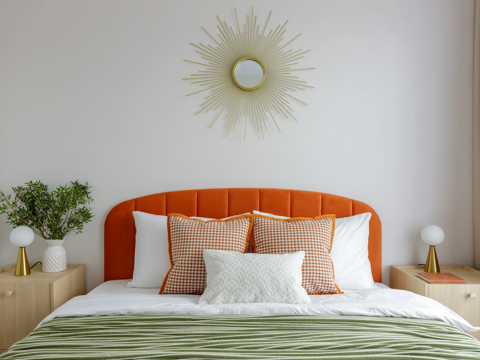 A bright bedroom with a burnt-orange headboard, a sunburst mirror and sage green bedding