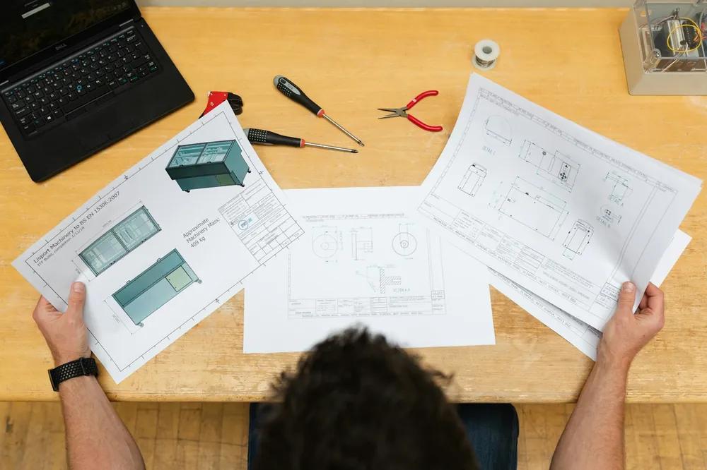 Someone reading printed floor plans and drawings on a wooden desk next to a laptop