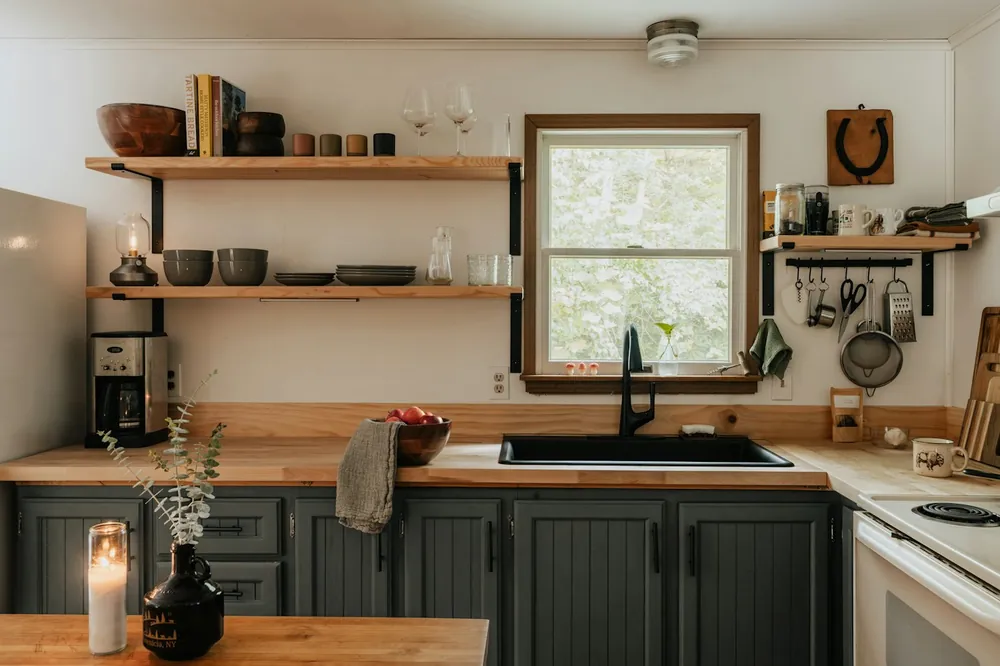 A kitchen with dark green units, open wooden shelves and a window over the sink