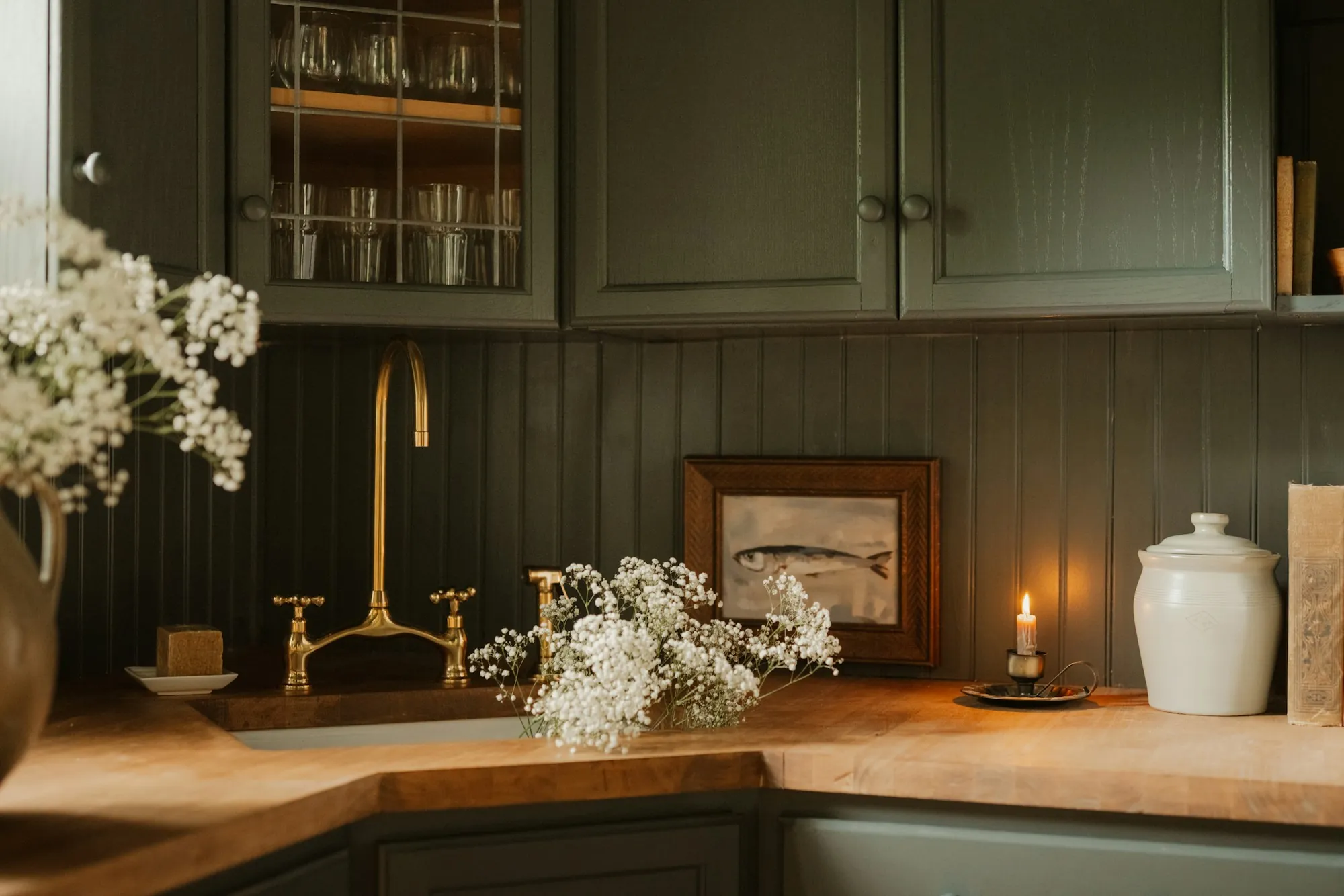 A dark sage kitchen with a brass tap, wooden worktops and white flowers