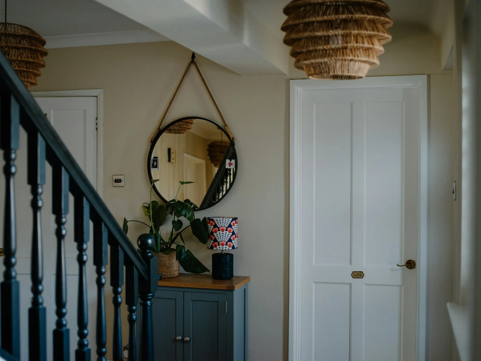 A hallway with a navy staircase, a round mirror and a woven pendant light
