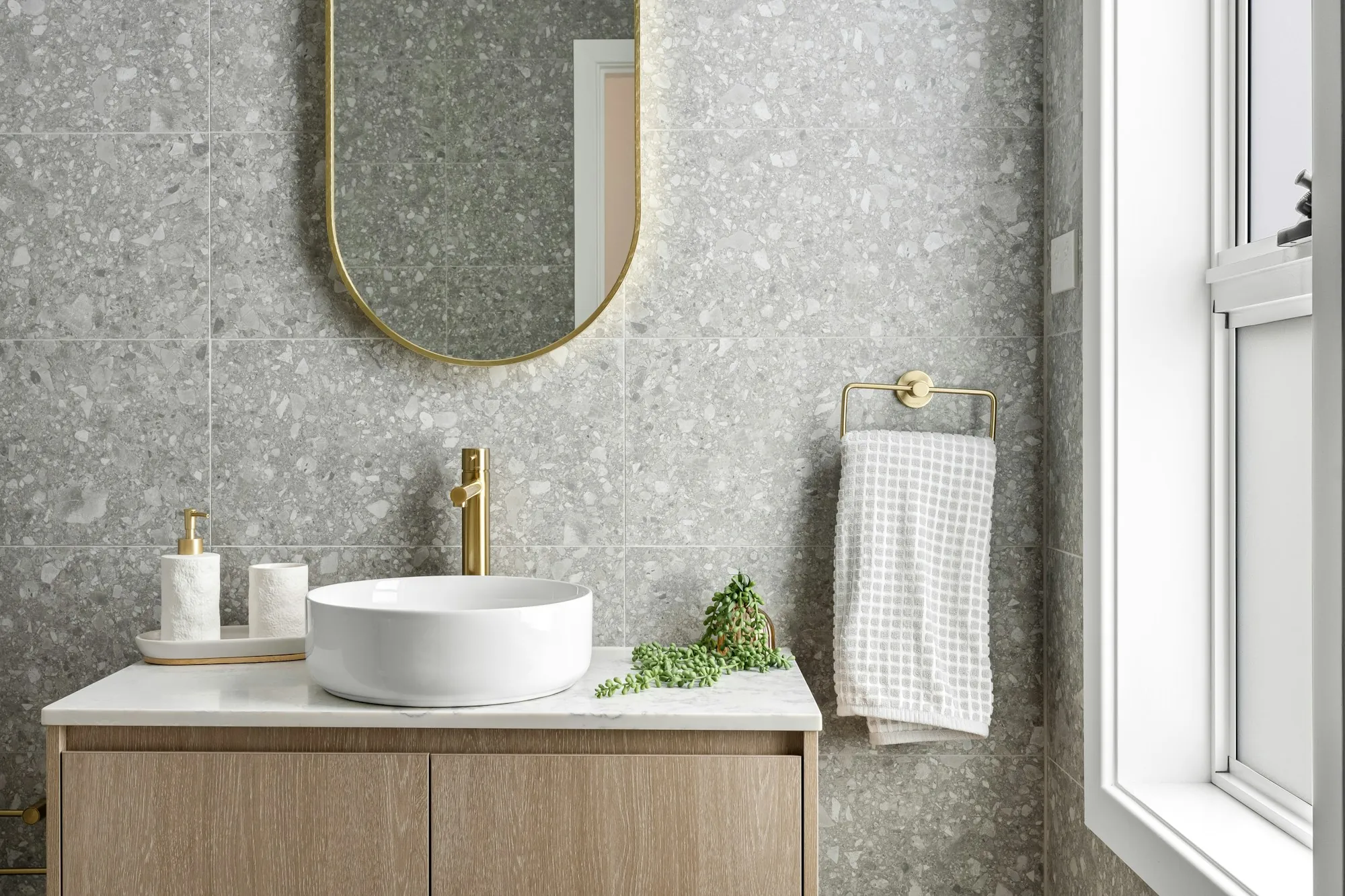 A bathroom with grey terrazzo walls, an arched brass mirror and a pale oak vanity