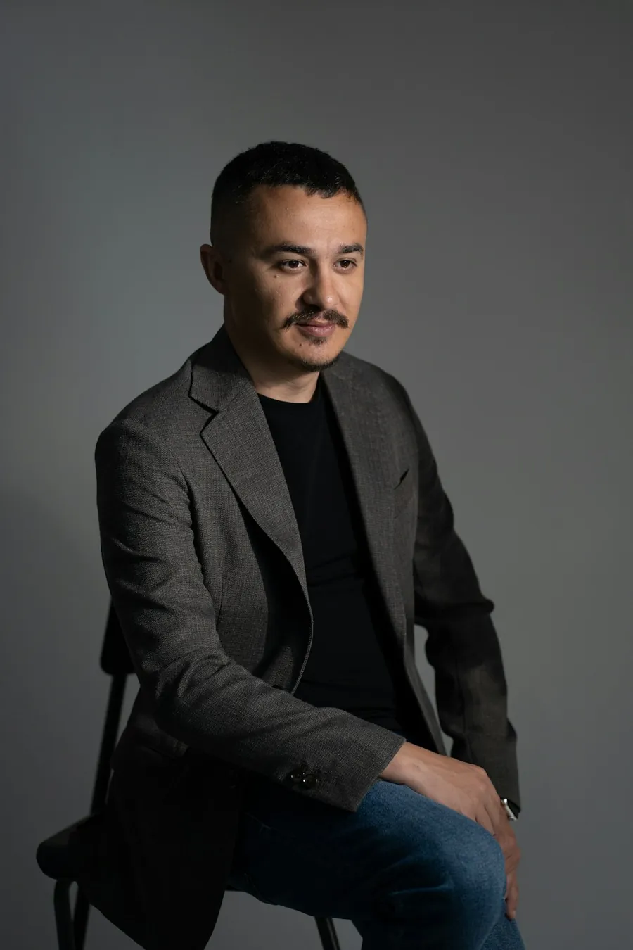 Sam, in a dark jacket, sitting on a chair against a grey wall