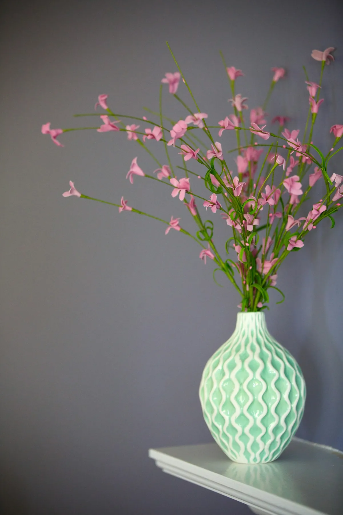 A green ceramic vase of pink blossom against a grey wall