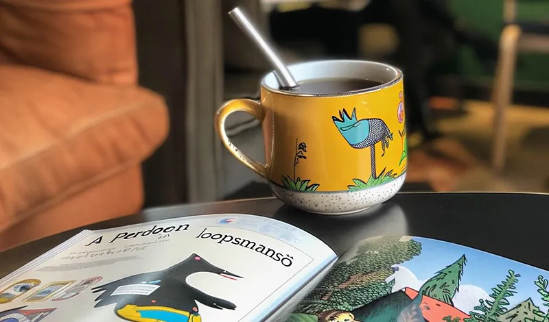 A yellow mug painted with a blue bird, beside an open book
