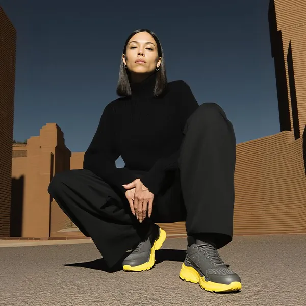 A woman in black crouching in yellow trainers in front of a brick building