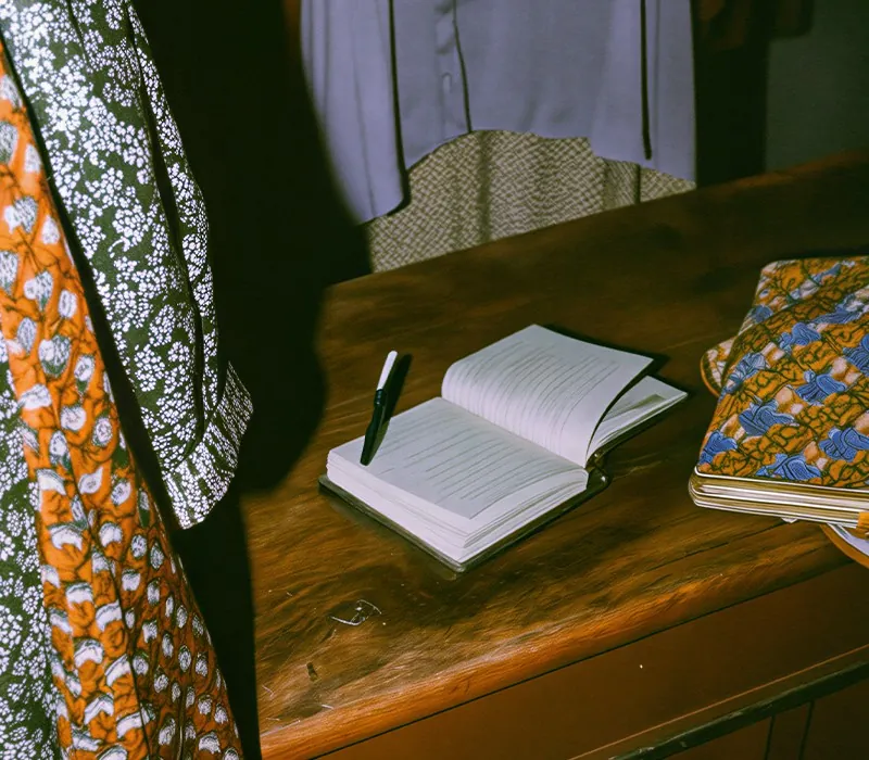 An open notebook and a pen on a wooden desk between patterned fabrics