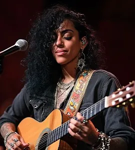 A woman with curly hair playing an acoustic guitar into a microphone