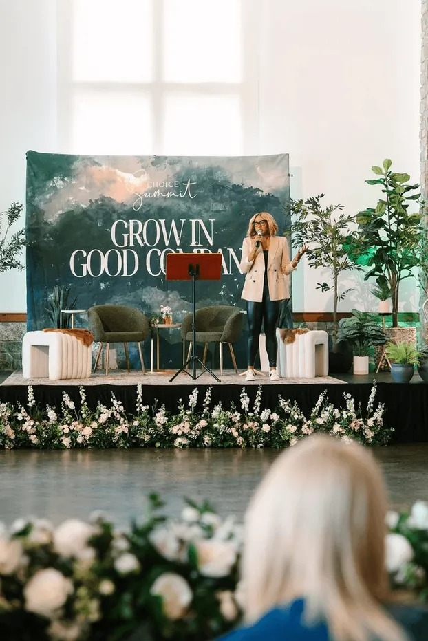 A speaker on a conference stage framed with white flowers