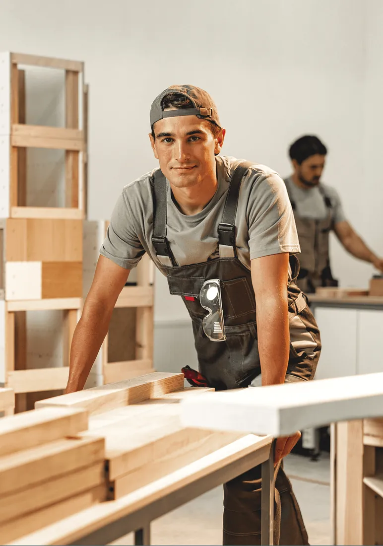 A young carpenter in overalls leaning on a workbench