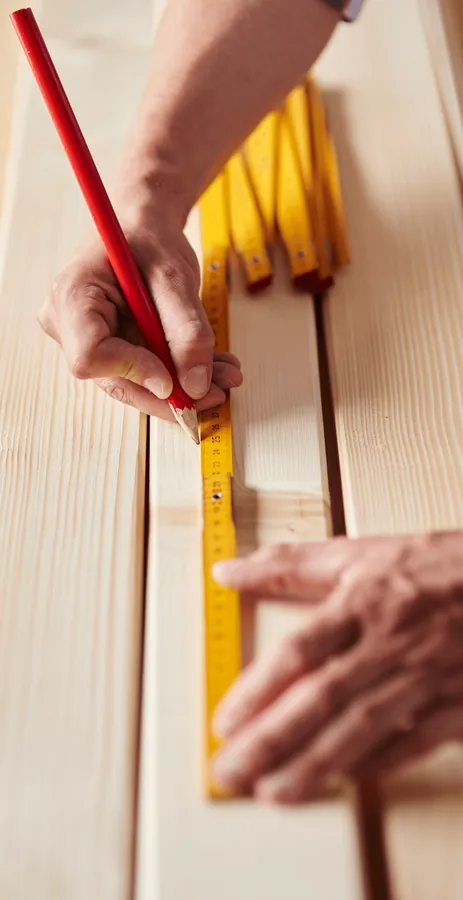 A hand marking a board with a red pencil and a yellow folding rule