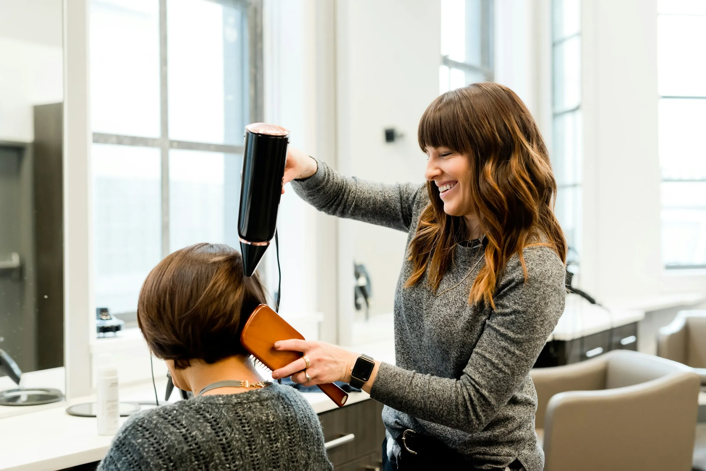 A stylist smiling as she blow-dries a client’s bob in a bright salon