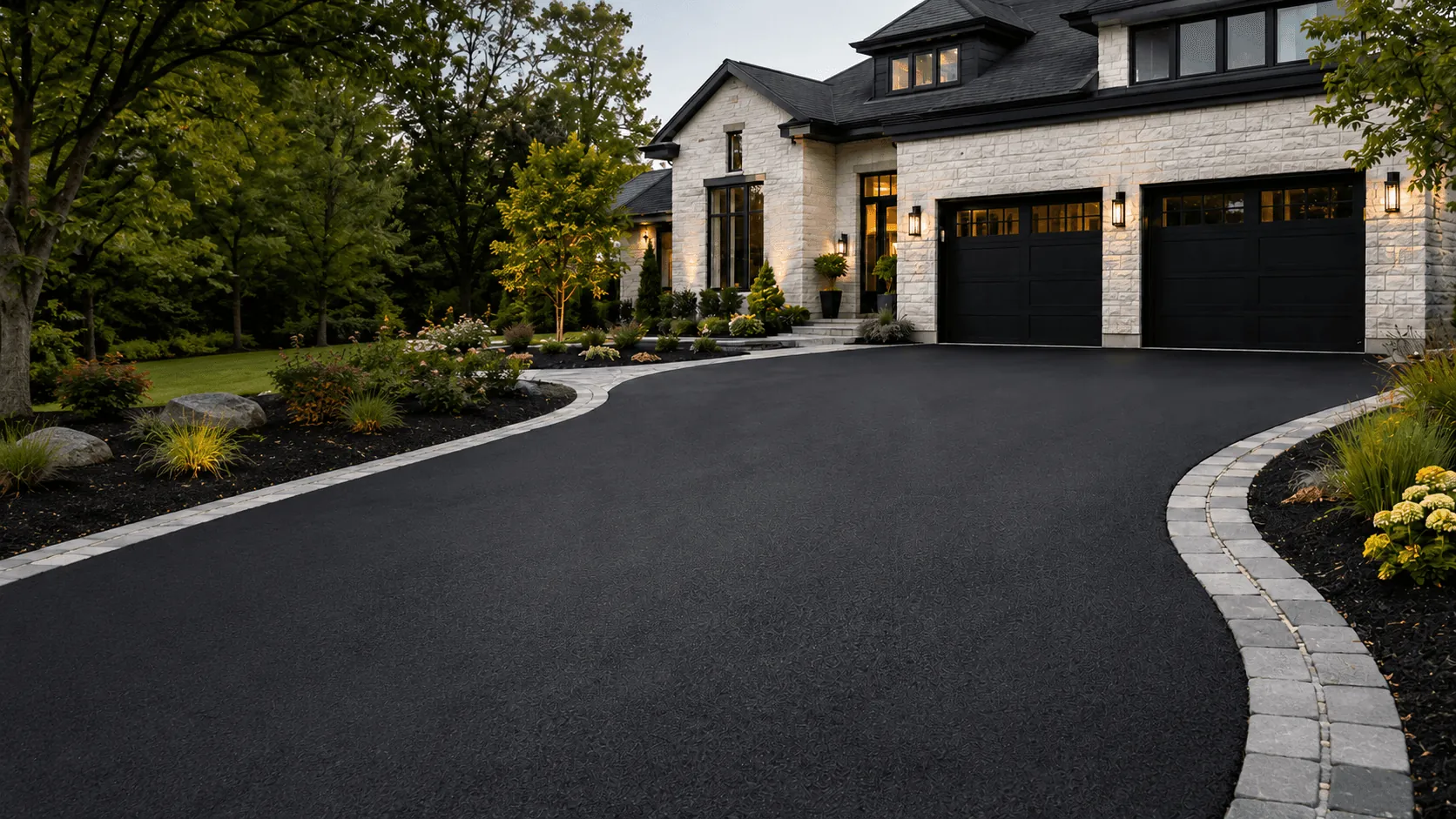 A freshly sealed black driveway curving up to a stone house at dusk