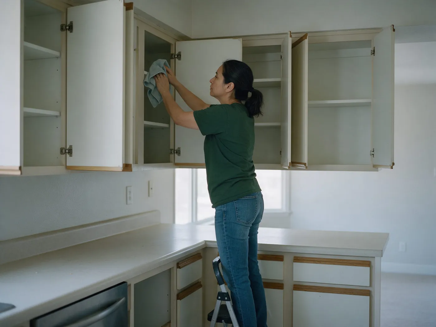 Cleaner on a step stool wiping the inside of empty kitchen cabinets