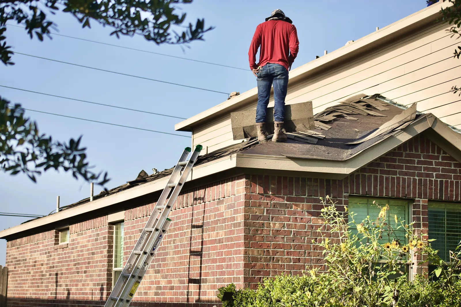 A tradesman on the roof of a brick house with a ladder against the eaves