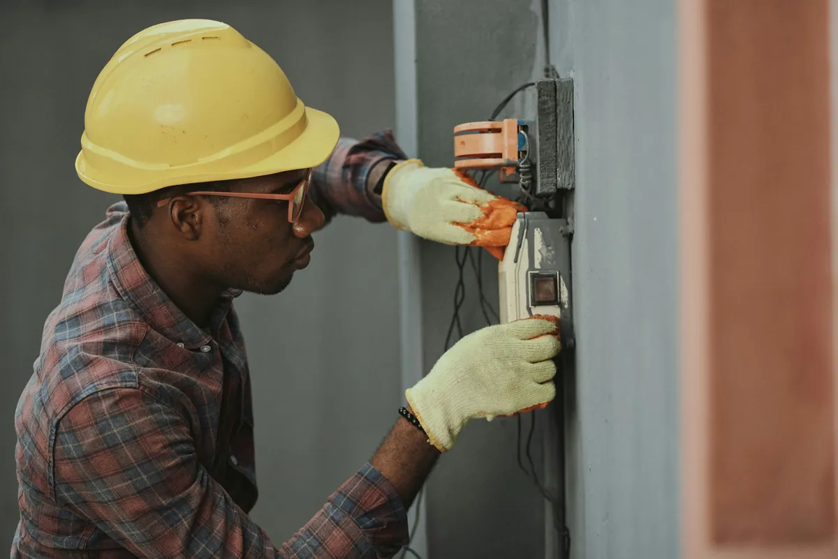An electrician in a yellow hard hat testing a wall panel