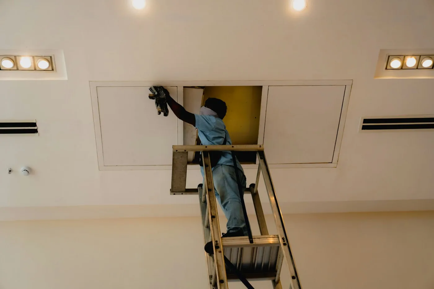 An electrician on a ladder wiring a light through a ceiling hatch